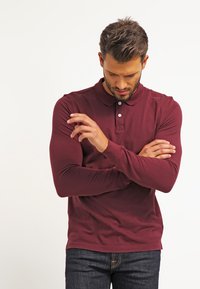 Camisa polo de manga larga burdeos con cuello, tapeta de tres botones y diseño ajustado, combinada con jeans oscuros.