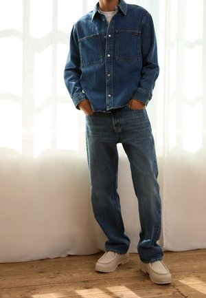 Personne portant une veste en jean bleu foncé et un jean, debout les mains dans les poches, avec des chaussures blanches sans lacets sur un sol en bois.