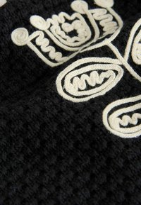 Lähikuva mustasta teksturoidusta kankaasta, jossa on kermaisen värinen kohokuvioitu kirjailu abstrakteilla, kaarevilla viivakuvioilla.