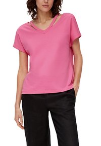 Różowa bluzka z krótkim rękawem i dekoltem w serek z unikalnymi wycięciami na ramiączkach. Posiada luźny krój i gładką strukturę materiału. Zestawiona z czarnymi spodniami.
