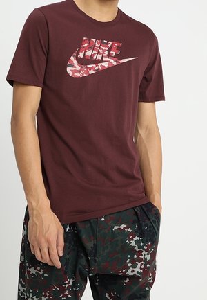 Mężczyzna w bordowej koszulce Nike z różowym logo w kamuflażu oraz spodniach w zielony, szary i czerwony wzór kamuflażu, stojący z jedną ręką w kieszeni.