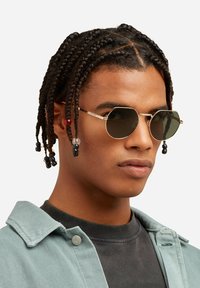 Ochelari de soare octogonali cu ton auriu și lentile întunecate. Modelul are părul împletit cu accente de mărgele, purtând o jachetă albastră deschisă peste un tricou negru.