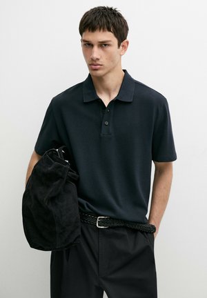 Junger Mann in dunklem Poloshirt und schwarzer Hose hält eine schwarze Jacke und steht vor schlichtem weißem Hintergrund mit neutralem Gesichtsausdruck.