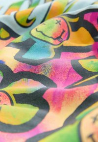 Bunt bedruckter Stoff mit einer Mischung aus grünen, pinken, blauen und orangenen abstrakten Mustern, umrandet von kräftigen schwarzen Linien und cartoonhaften Gesichtern.
