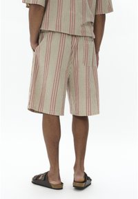 Pantaloni scurți cu dungi bej, roșii și albe, având un buzunar la spate. Purtați cu sandale închise la culoare, evidențiind un fit relaxat și un design casual.