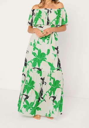 Kobieta ubrana w długą suknię odsłaniającą ramiona, z dużym zielono-czarnym kwiatowym wzorem, splatająca ręce na talii, widoczne bose stopy.