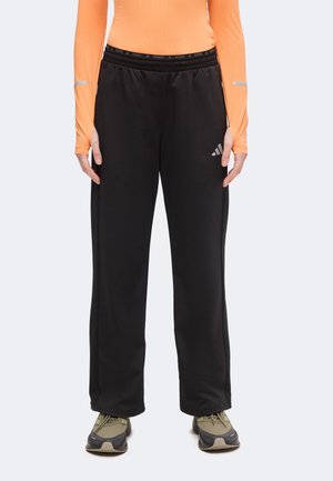 Personne portant un pantalon de survêtement Adidas noir, un haut orange à manches longues et des baskets vertes et grises, debout droit sur un fond uni.
