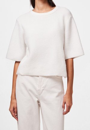 Vrouw draagt een loszittende witte gebreide trui met korte mouwen en een witte broek met hoge taille tegen een effen achtergrond.