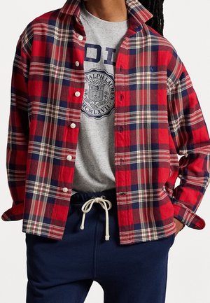 Person trägt ein rot-navy kariertes Hemd mit Knöpfen über einem grauen T-Shirt mit Ralph Lauren Logo und navyblaue Hosen mit Kordelzug.