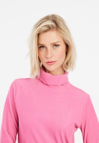 Rausvai dryžuotas turtleneck megztinis su prigludusia forma. Pabrėžia vertikalias juostas ir aukštą apykaklę, pagamintas iš minkštos, tekstūruotos medžiagos.