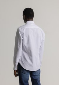 Hombre de espaldas vistiendo una camiseta blanca de manga larga y jeans azules, frente a un fondo claro y sencillo.