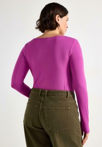 Långärmad topp i lysande rosa tyg, med rundad halsringning, kombinerad med olivgröna högmidjade byxor med bakfickor.