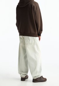 Felpa marrone con vestibilità ampia, abbinata a jeans baggy verde chiaro. Sneakers scure con superficie testurizzata e accenti a contrasto.