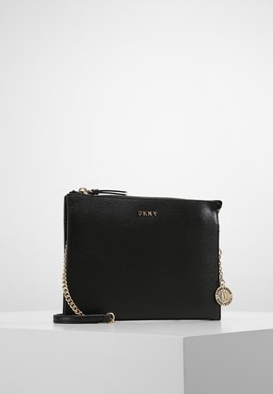 Black DKNY leather crossbody bag with gold chain strap and round Donna Karan logo charm on white surface background.