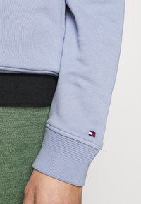 Sudadera de color azul claro con puños acanalados, superpuesta sobre una tela texturizada de color verde oscuro. Un pequeño logo muestra colores rojo, blanco y azul marino.
