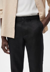Black linen trousers with a smooth texture, featuring a button closure and side pockets, paired with a light beige button-up shirt.