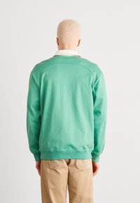 Grön sweatshirt med långa ärmar, ribbade muddar och fåll, med en enkel design och mjuk textur. Bärs tillsammans med beige kordflorbyxor.