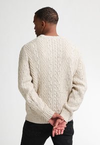 Ljus beige stickad tröja med en strukturerad kabelmönster, rund halsringning och ribbade ärmslut och fåll. Stylad med svarta byxor.