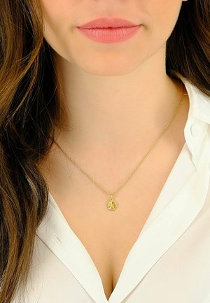 Goldkette mit einer zarten Kette und einem runden Anhänger mit filigranen Designdetails, getragen über einem weißen Hemd mit Kragen.