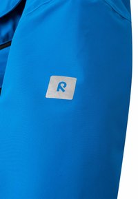 Giacca impermeabile blu con una texture liscia e una toppa con logo argentato sulla parte superiore del braccio, caratterizzata da un design ad alta visibilità.