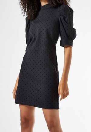 Czarna sukienka do kolan z bufkami na krótkich rękawach i małym wzorem w groszki, noszona przez osobę o kręconych włosach.