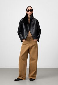 Giacca di pelle nera con colletto in pelliccia sintetica, abbinata a pantaloni larghi beige e scarpe ballerina nere, su uno sfondo neutro.