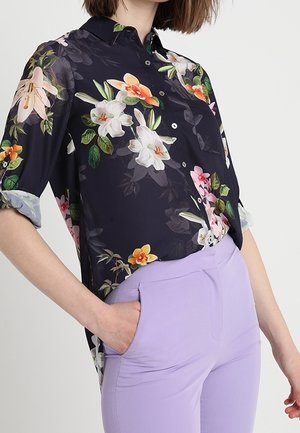 Nainen, jolla on yllään tumma kukallinen napillinen paita rullatuilla hihoilla ja vaaleanliilat istuvat housut, toinen käsi taskussa.