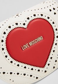 Bílá kožená kabelka s červeným srdcem uprostřed a logem "LOVE MOSCHINO". Lemovaná perforovanými srdíčky a cikcak stehy pro texturu.