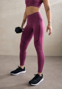 Paarse sportlegging met een gladde textuur en contrasterende zijkleurpatronen, gecombineerd met zwarte sneakers en een zwarte dumbbell vastgehouden.