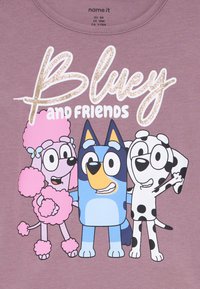 Lilla skjorte med trykk av tegneseriefigurer, Bluey, en blå hund, og to venner, en rosa puddel og en hvit hund med svarte flekker.
