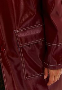 Hnedý kožený kabát zblízka so širokou kapsou, viditeľným šitím a okrúhlym gombíkom na látke. Hladká, lesklá textura.