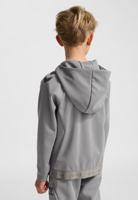 Grå hættetrøje med en tekstureret finish og snøre i hætten, med kontrasterende mørke detaljer på skuldrene og et bånd med logo ved taljen.