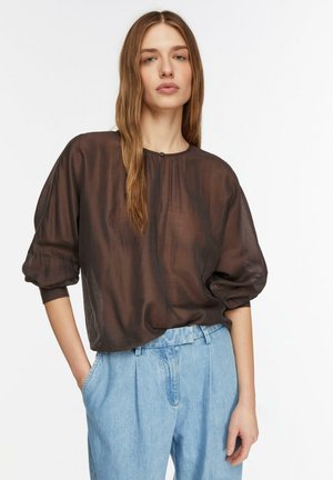 Młoda kobieta ubrana w przezroczystą brązową bluzkę z marszczonymi rękawami i jasnoniebieskie dżinsy z wysokim stanem, stojąca z jedną ręką w kieszeni.