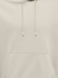 Vaaleanbeige huppari, jossa on kiristysnyörit ja etupuolen kengurutasku näkyvine ompeleineen.