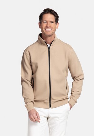 Man die lacht en een beige jack met zwarte ritsdetails en witte broek draagt, tegen een eenvoudige lichte achtergrond.