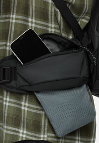 Zwarte stoffen heuptas met een ritsvak waarin een smartphone ligt. Een grijs, gestructureerd voorwerp is gedeeltelijk zichtbaar onder de tas. Achtergrond van een groen geruit shirt.