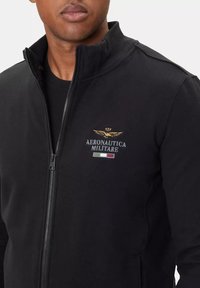 Giacca nera con zip realizzata in tessuto morbido, caratterizzata da un logo ricamato con ali e colori della bandiera italiana sul lato sinistro del petto.