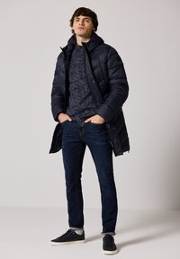 Námořnická pufovaná bunda, vzorovaná tmavá košile, tmavé džíny a černé tenisky. Bunda má kapuci a viditelné švy.