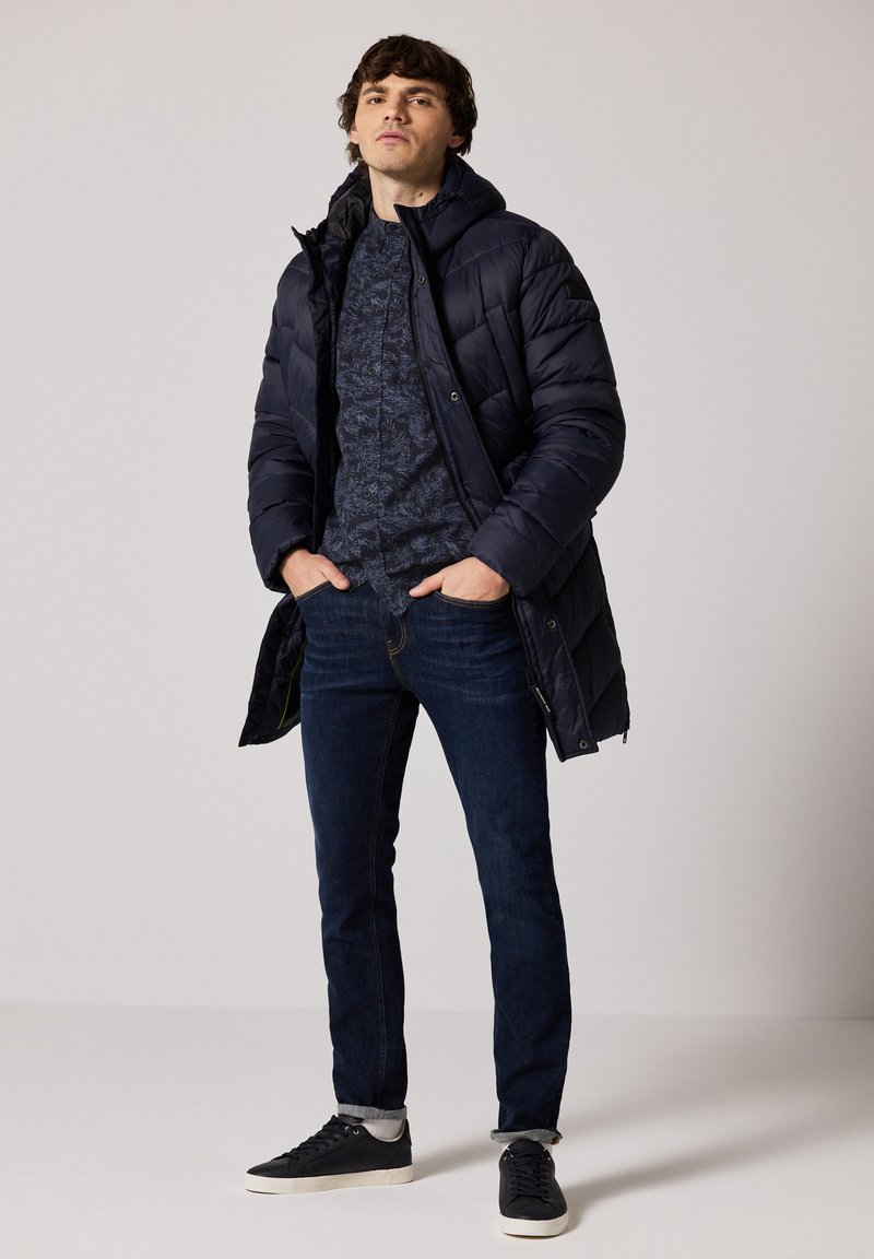 Námořnická pufovaná bunda, vzorovaná tmavá košile, tmavé džíny a černé tenisky. Bunda má kapuci a viditelné švy.