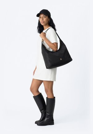 Woman wearing white short dress, black cap, and knee-high black boots, holding large black shoulder bag with gold logo.