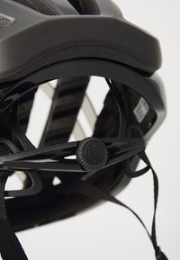 Svart sykkelhjelm med en teksturert ytterskall, justerbart belte med en rund dial, og en innside med ventilasjonsåpninger.