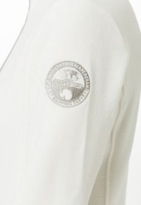 Manga de chaqueta de forro polar blanca con parche bordado circular que muestra un mapa del mundo y texto que incluye "Mapping Survey" en el brazo superior.