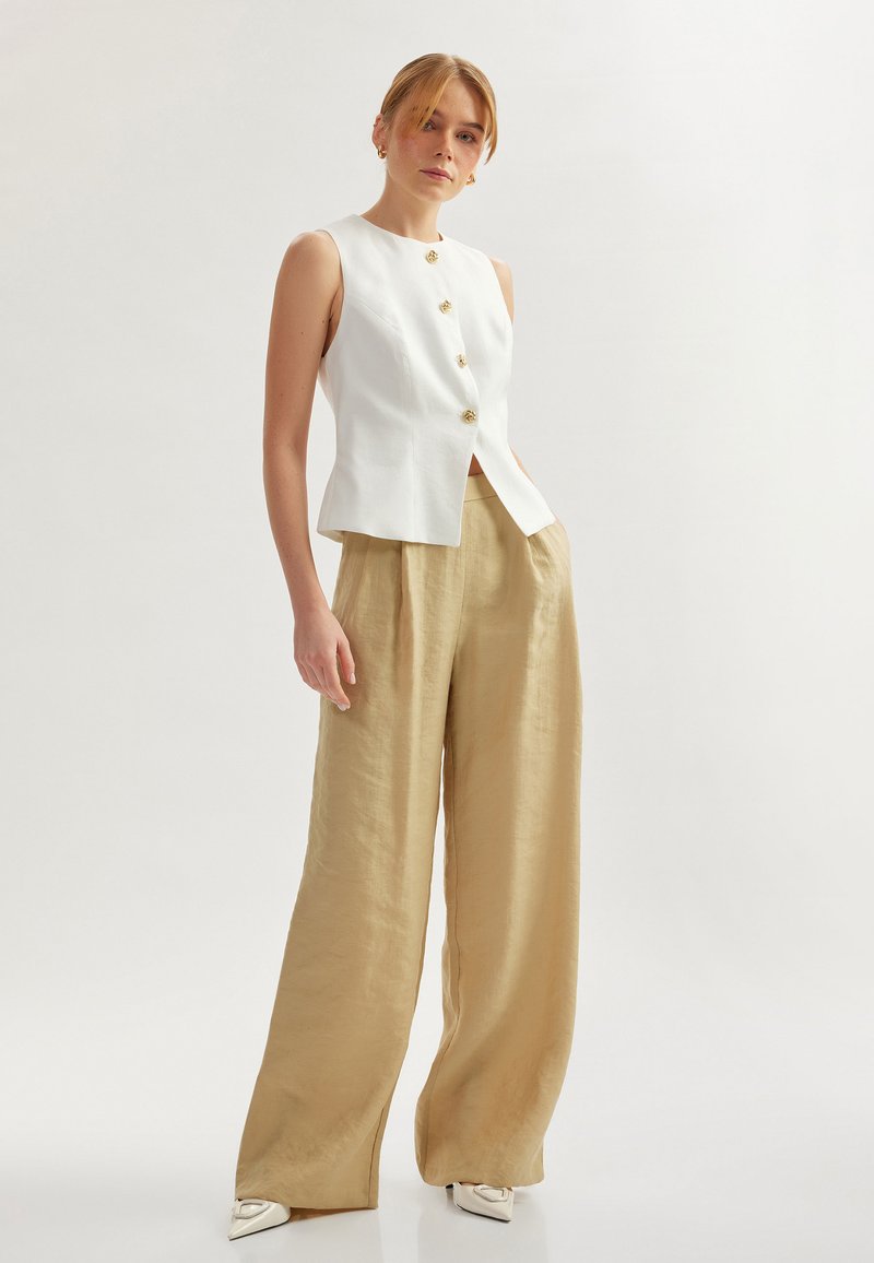 Blusa bianca senza maniche con bottoni dorati e orlo dritto, abbinata a pantaloni larghi in lino beige, caratterizzati da una consistenza liscia.
