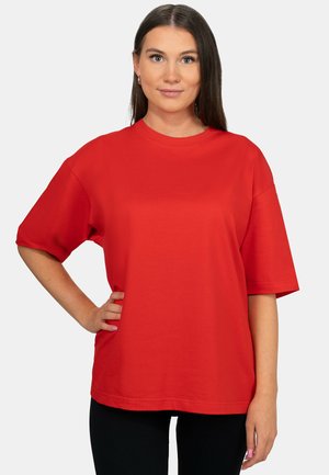 Vrouw met lang bruin haar, die een losse rode korte mouwen blouse en zwarte broek draagt, staat met één hand op haar heup tegen een effen achtergrond.