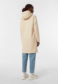 Lungo cappotto beige con cappuccio trapuntato, abbinato a jeans azzurri con risvolti e sneakers bianche. Texture liscia, design minimal e nessun motivo visibile.