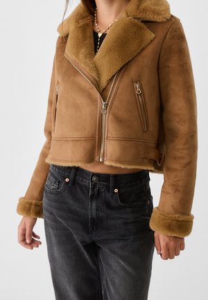Vrouw draagt een korte bruine shearling jas met brede kraag en ritszakken over zwarte high-waisted jeans.