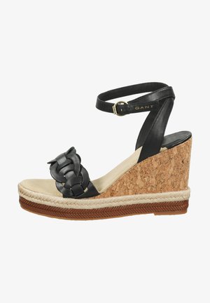 Black leather wedge sandals with a braided strap detail, cork wedge heel, and a natural jute band around the sole. Gold buckle accent.