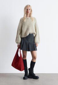 Flauschiger Cremepullover, grauer plissierter Rock, schwarze kniehoge Socken und schwarze Plateaustiefel. Hält eine rote Wildleder-Handtasche.