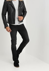 Uomo con giacca di pelle nera con colletto di pelliccia, camicia a righe grigie, jeans neri e stivali in camoscio neri, in piedi con una gamba incrociata.