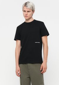 Zwarte katoenen t-shirt met korte mouwen, ronde halslijn en klein wit logo aan de linkerzijde. Gepaard met groene cargobroek.